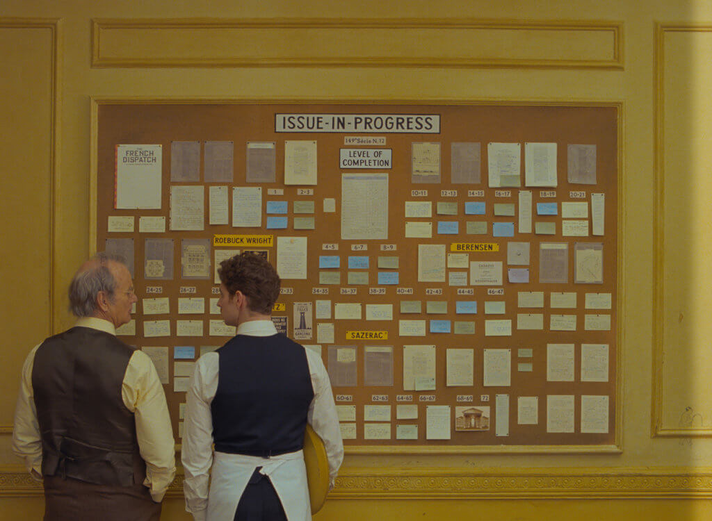 Still frame from The French Dispatch of characters looking at a community pin board